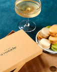 Coffret de Noël doré Atelier St Eustache à côté d'une assiette de macarons et d'une coupe de champagne, dans une ambiance festive et lumineuse pour Noël