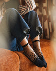 Chaussettes transparentes à pois noirs, portées par une femme assise sur un canapé cocooning en jean bleu