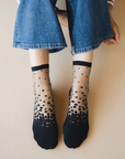 Chaussettes transparentes à pois noirs, portées par une femme assise par terre et portant un jean bleu