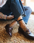 Chaussettes transparentes à pois noirs, portées avec des mocassins bordeaux et dorés sur un tapis moderne marron