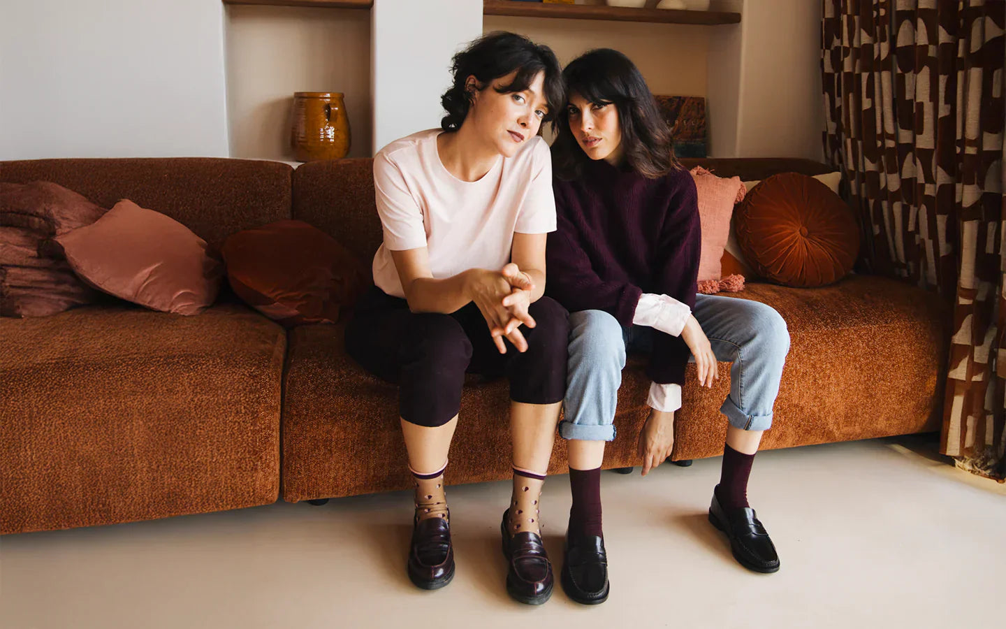 Deux femmes brunes assises sur un canapé vintage brun, l’une portant des chaussettes transparentes à petits cœurs rose clair et bordeaux, l’autre des chaussettes en coton bordeaux, toutes deux regardant l’objectif.