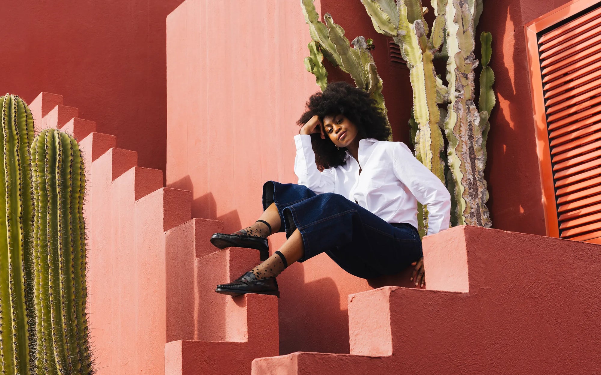 Mannequin portant des chaussettes transparentes noires à pois dégradés avec des mocassins noirs, assise sur des escaliers en béton rouge-ocre. Elle porte une chemise blanche et un pantalon noir.
