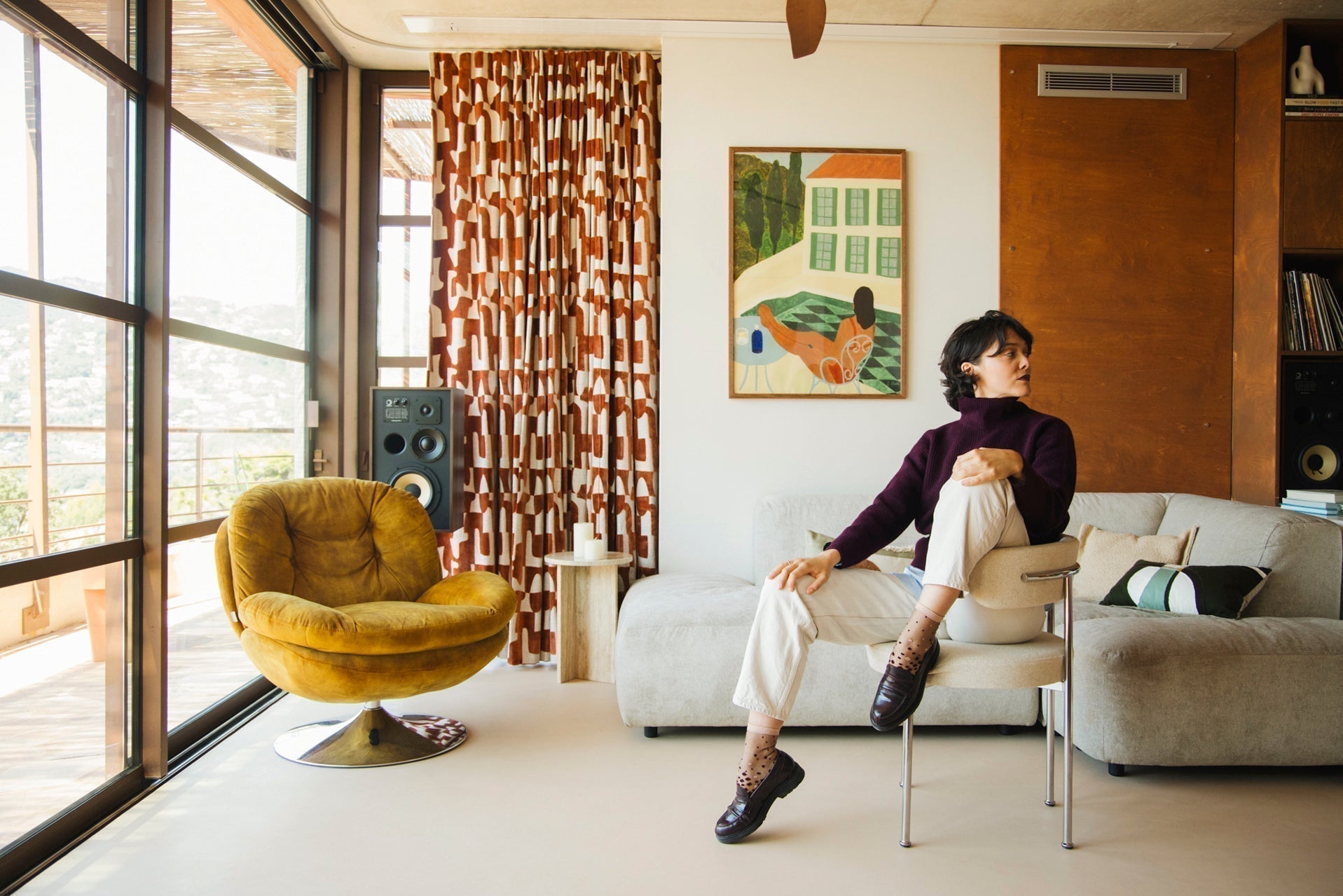 Mannequin qui porte des chaussettes transparentes au dégradé de poids bordeaux et doré, vêtue d'un pantalon beige, pull bordeaux et de mocassins noirs. Elle est assise sur une chaise beige, dans un salon au style rétro année 70, et regarde vers la droite.