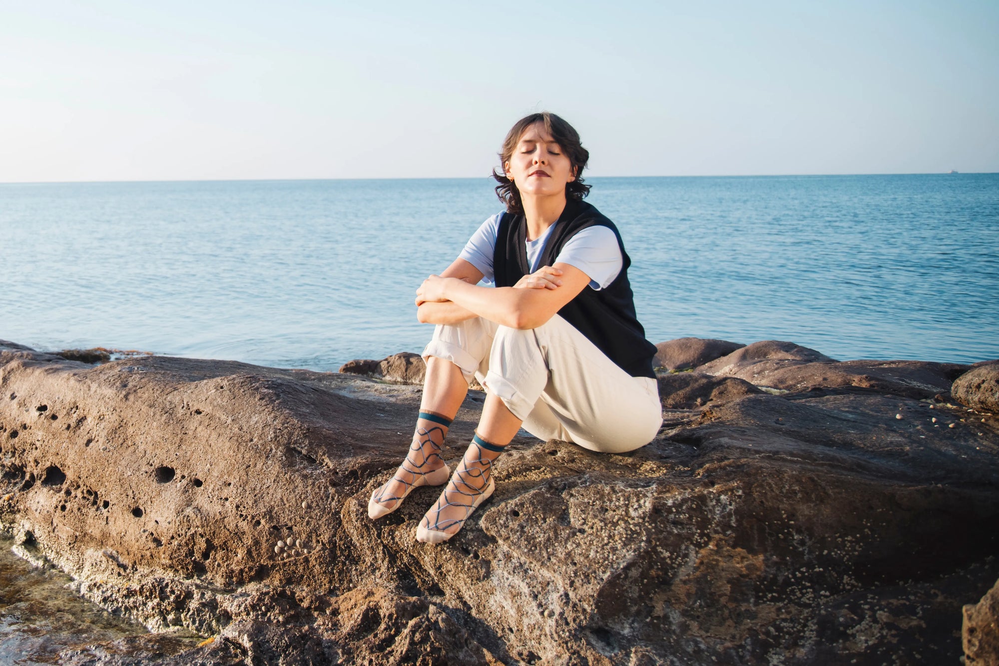 Femme portant des chaussettes transparentes aux courbes bleues, vêtue d'un pantalon crème, d'un tee-shirt bleu et d'une veste sans manche bleue foncé, assise sur des rochers au bord de la mer.