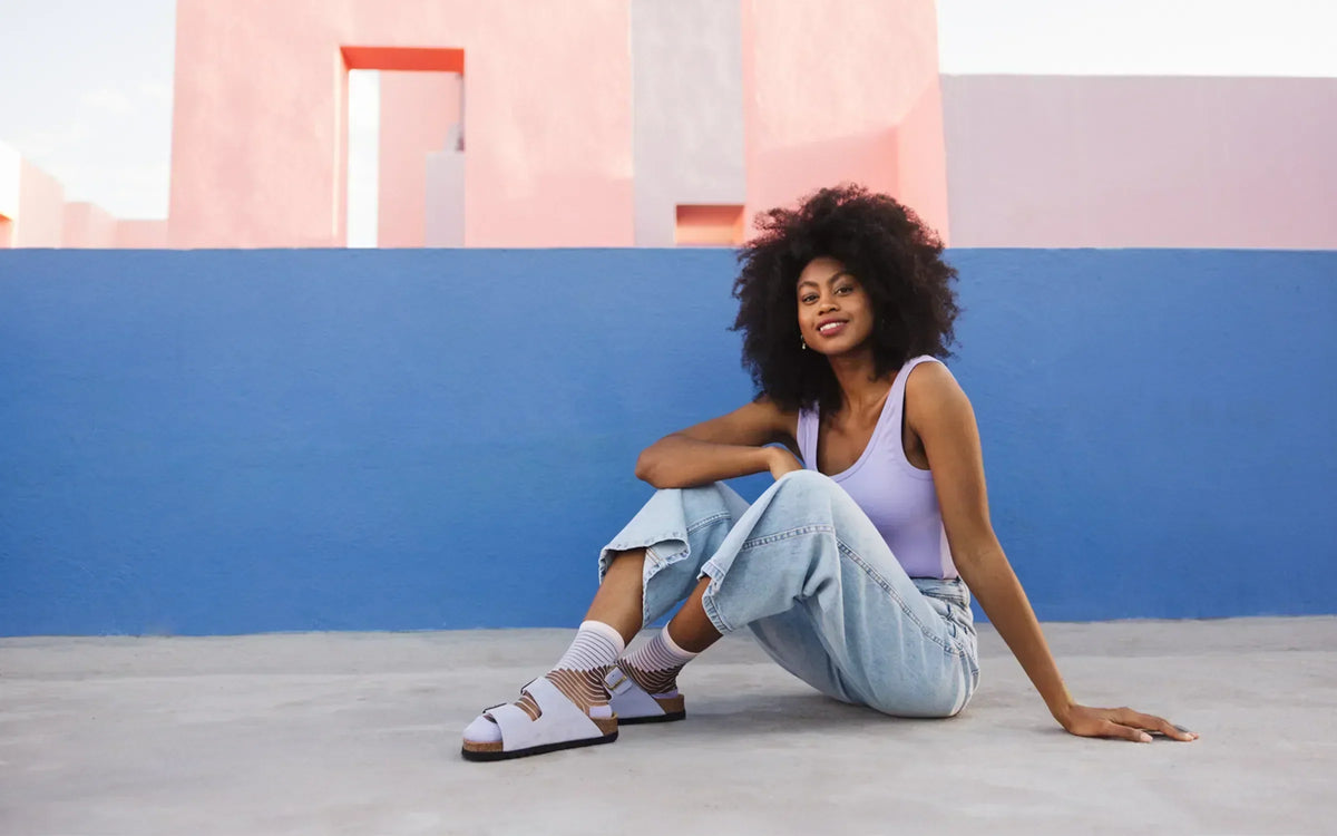 La photographie présente une mannequin assise de profil sur un sol en béton, dans un environnement extérieur devant un mur bleu. Elle porte des chaussettes transparentes ornées de pompons pailletés lilas, associées à des sandales ouvertes.