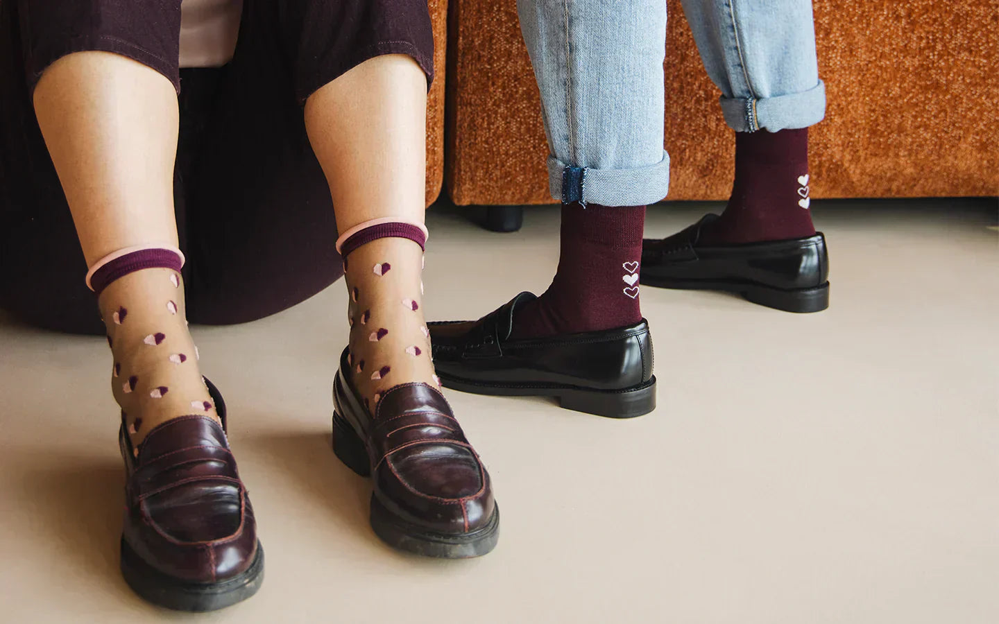 Zoom sur les pieds de deux mannequins : l’une porte des chaussettes transparentes ornées de petits cœurs. L’autre, porte des chaussettes bordeaux décorées de trois cœurs blancs alignés verticalement à l’arrière de chaque cheville.