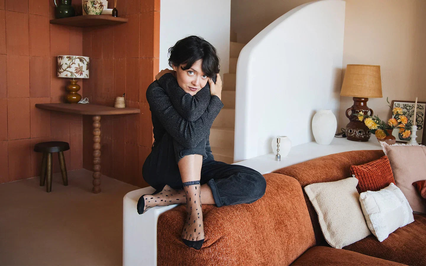 Plan large sur une femme assise sur le rebord d’un canapé, se tenant les bras autour du corps. Elle porte une tenue noire et des chaussettes transparentes pailletées noir et argent.