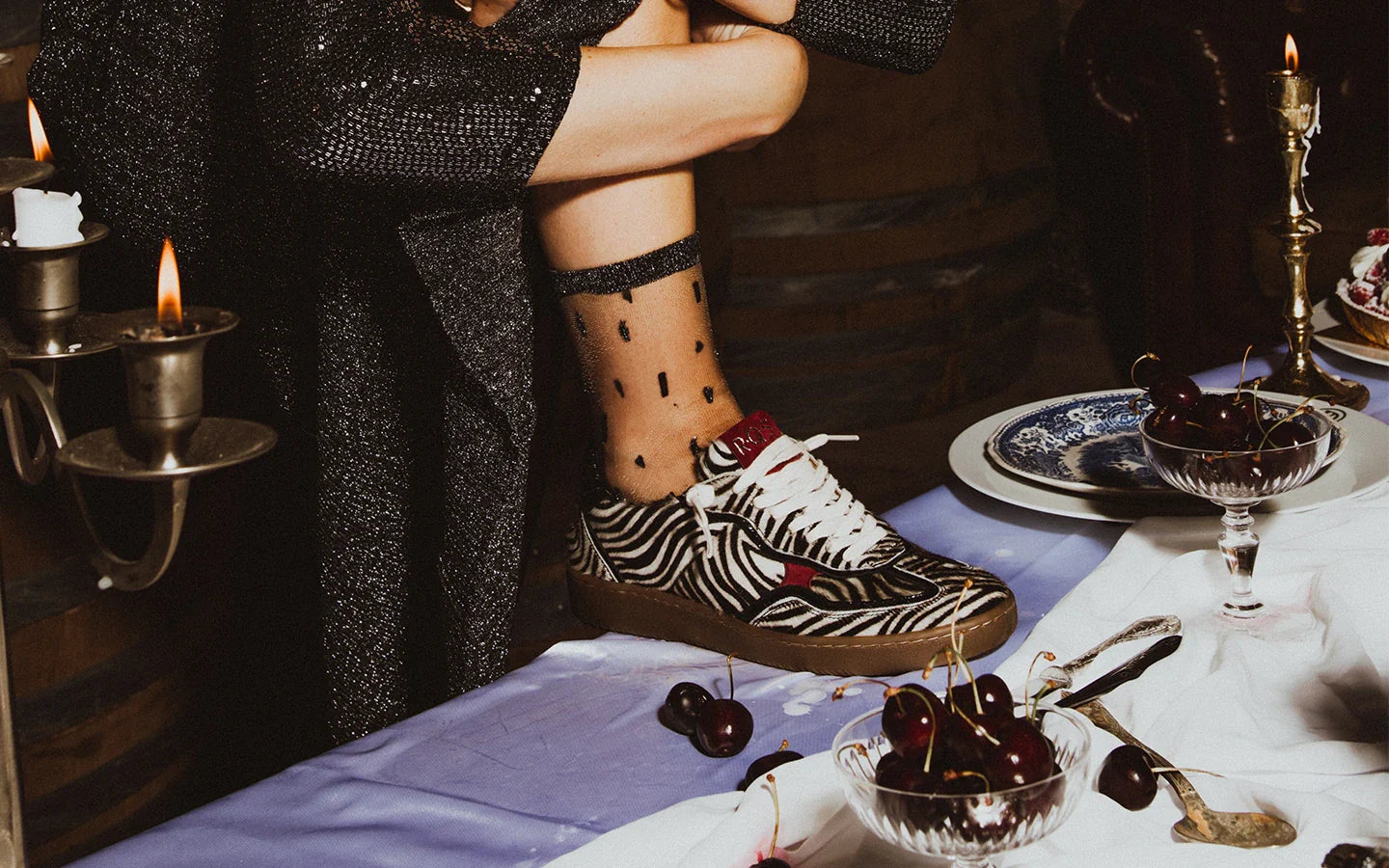 Zoom sur le pied d’une femme portant des chaussettes transparentes à motifs rectangulaires verticaux pailletés noir et argent. Elle porte des chaussures zébrées, et son pied repose sur une table recouverte d’une nappe blanche.