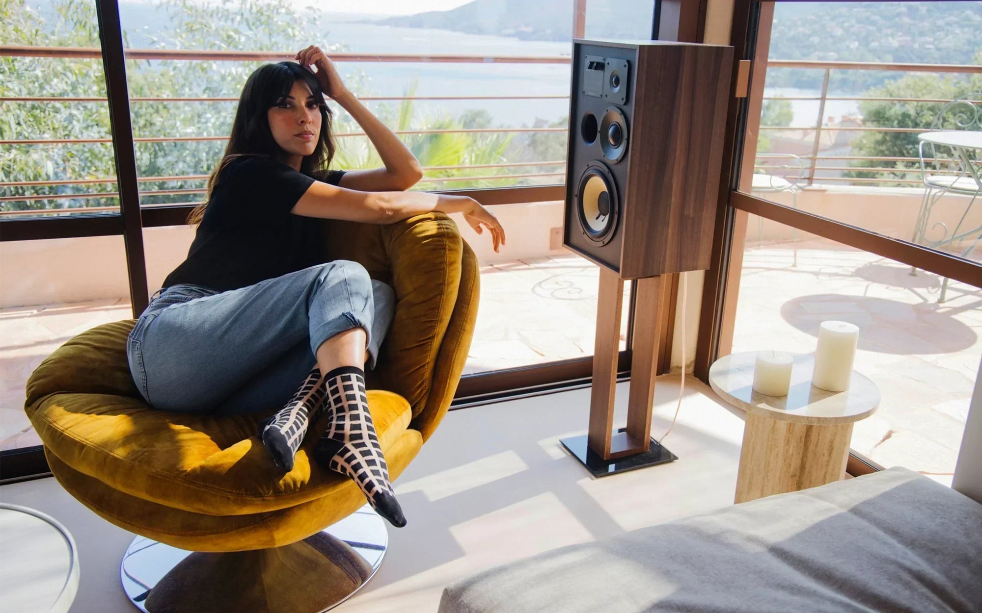 C’est une photographie d’une femme assise sur un fauteuil jaune moutarde. Elle porte des chaussettes transparentes architecturales à carreaux noirs.