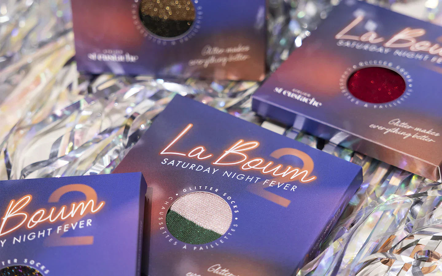 Zoom sur quatre boîtes carrées de chaussettes à paillettes de la marque Saint Eustache. Les packagings, de couleurs bleu, violet, rose et orange, sont disposés au sol sur un rideau à paillettes argentées qui sert de fond.