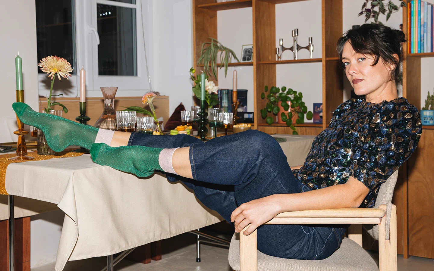 Une femme brune est assise sur une chaise, posant ses pieds sur une table décorée pour les fêtes, portant un haut pailleté bleu, rose et vert et des chaussettes vert sapin à paillettes avec bord roulotté rose poudré.
