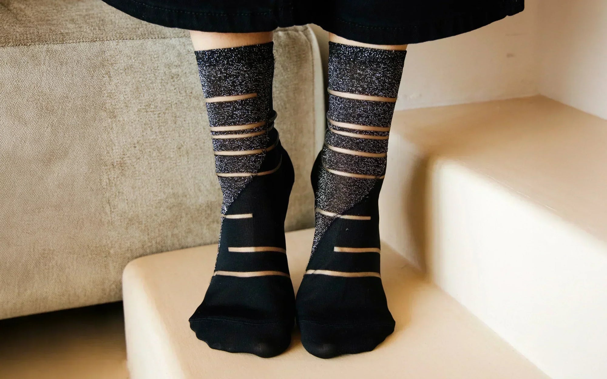 Gros plan sur les pieds d’une femme portant des chaussettes transparentes noires et argentées à motifs géométriques. Elle porte un pantalon noir et se tient sur la pointe des pieds, posée sur la première marche d’un escalier beige