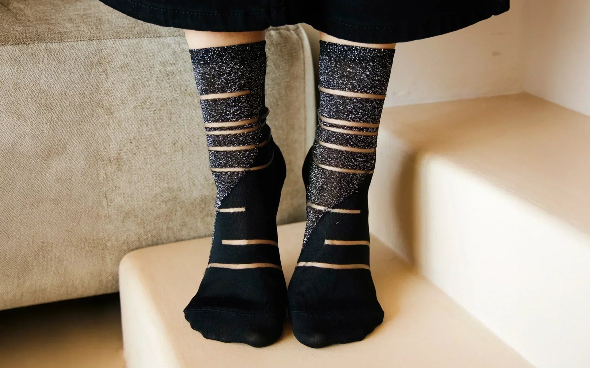 Gros plan sur les pieds d’une femme portant des chaussettes transparentes noires et argentées à motifs géométriques. Elle porte un pantalon noir et se tient sur la pointe des pieds, posée sur la première marche d’un escalier beige