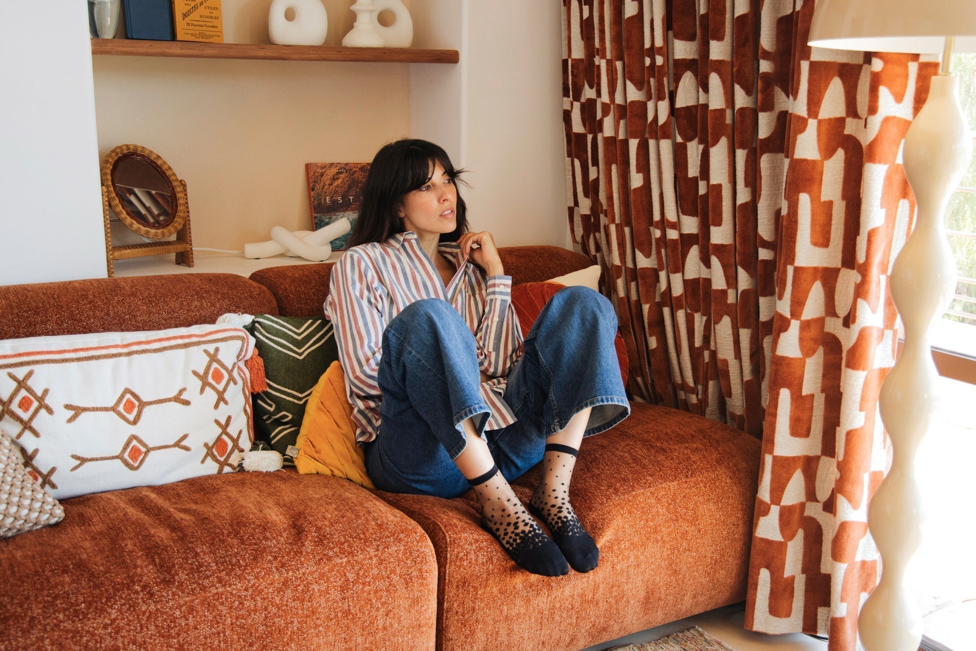 Femme qui porte des chaussettes transparentes au dégradé de poids noirs, vêtue d'une chemise rayée rouge et bleue et d'un jean bleu. Elle est assise sur un canapé couleur brique dans un salon au style année 70, regardant au loin.