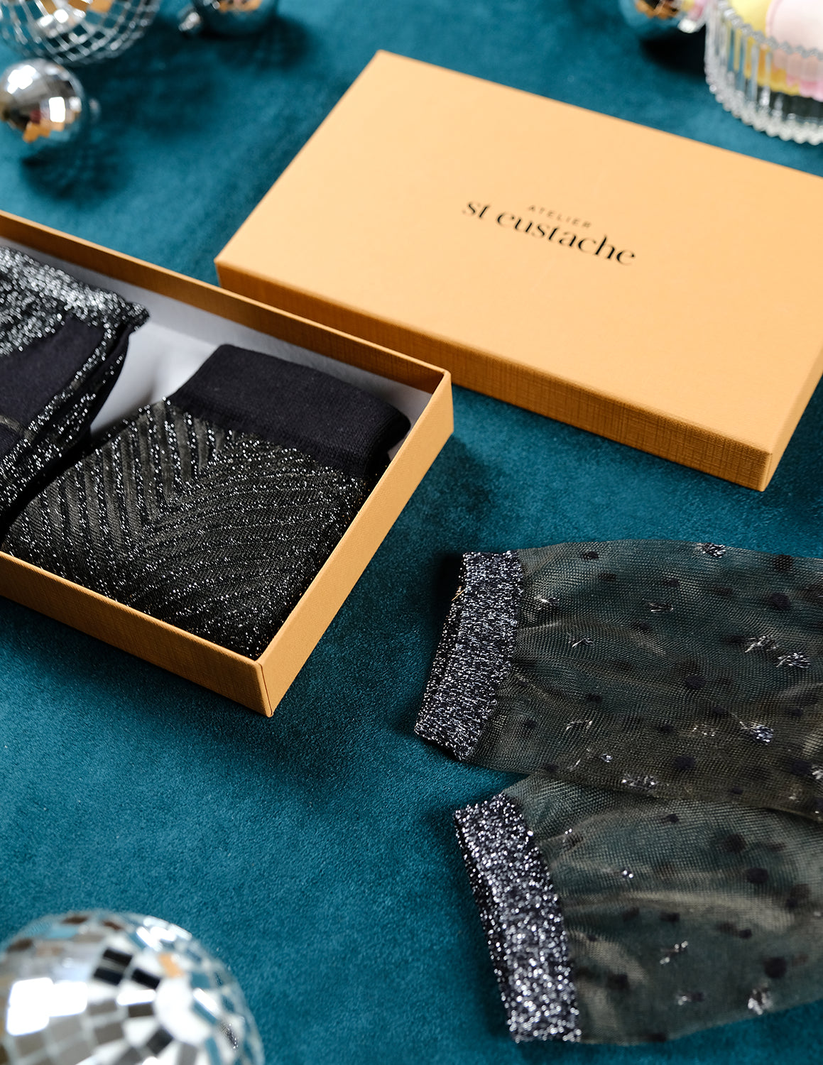 Vue de côté du coffret de Noël argenté Atelier St Eustache avec la paire de chaussettes transparentes Nakameguro silver posée à plat et les modèles Sapin Silver et Tate Modern Silver pliés, dans une ambiance hivernale pour Noël