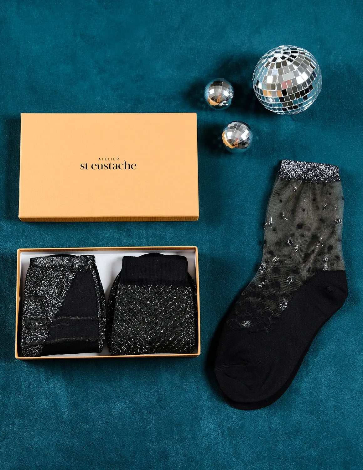 Coffret de Noël argenté Atelier St Eustache avec la paire de chaussettes transparentes à paillettes Nakameguro silver posée à plat et les modèles Sapin Silver et Tate Modern Silver pliés, dans une ambiance hivernale pour Noël