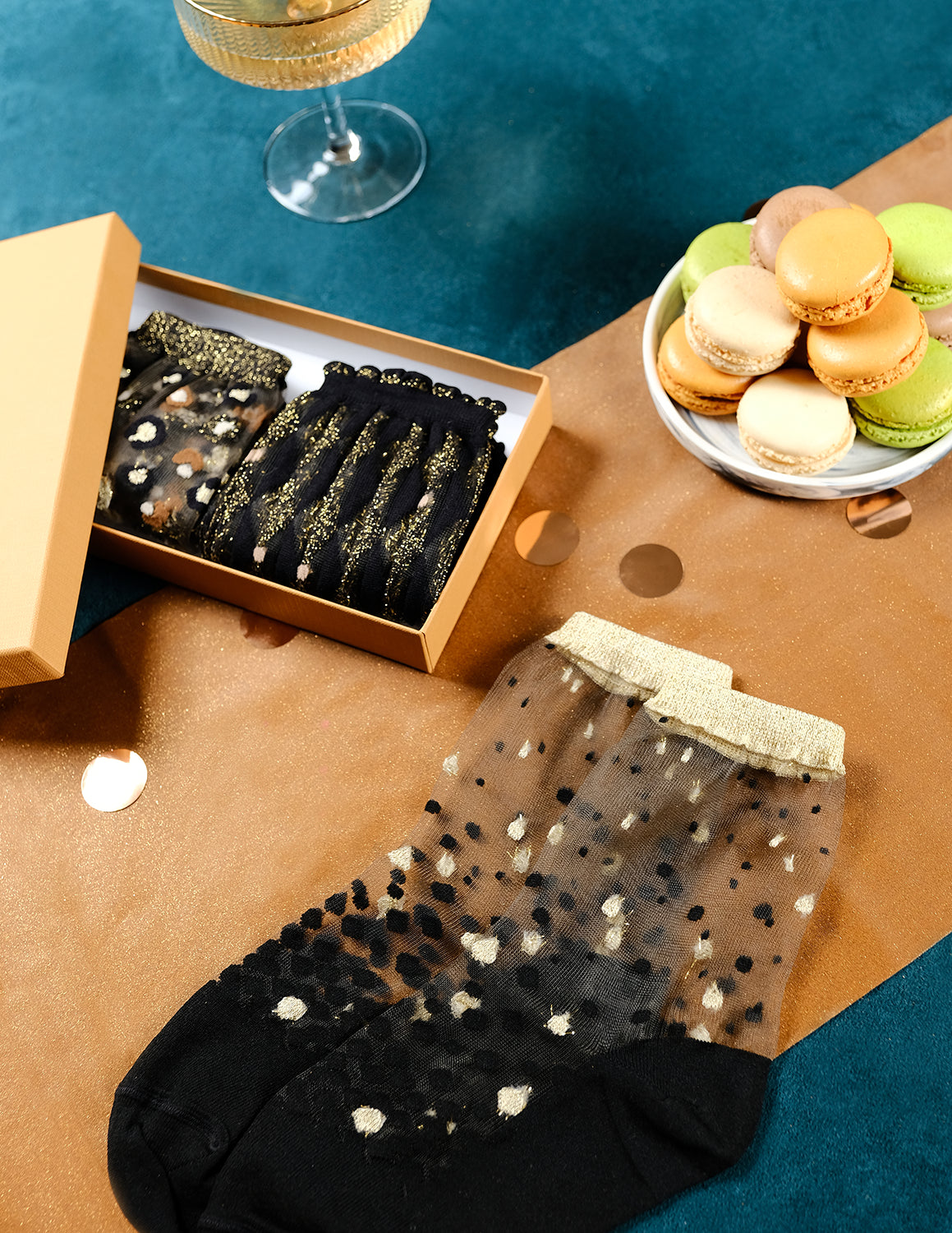 Coffret de Noël doré Atelier St Eustache avec la paire de chaussettes transparentes à paillettes Nakameguro Or posée à plat et les modèles Arlecchino dorée et Horseshoe Bend Gold pliés, dans une ambiance festive et lumineuse pour Noël