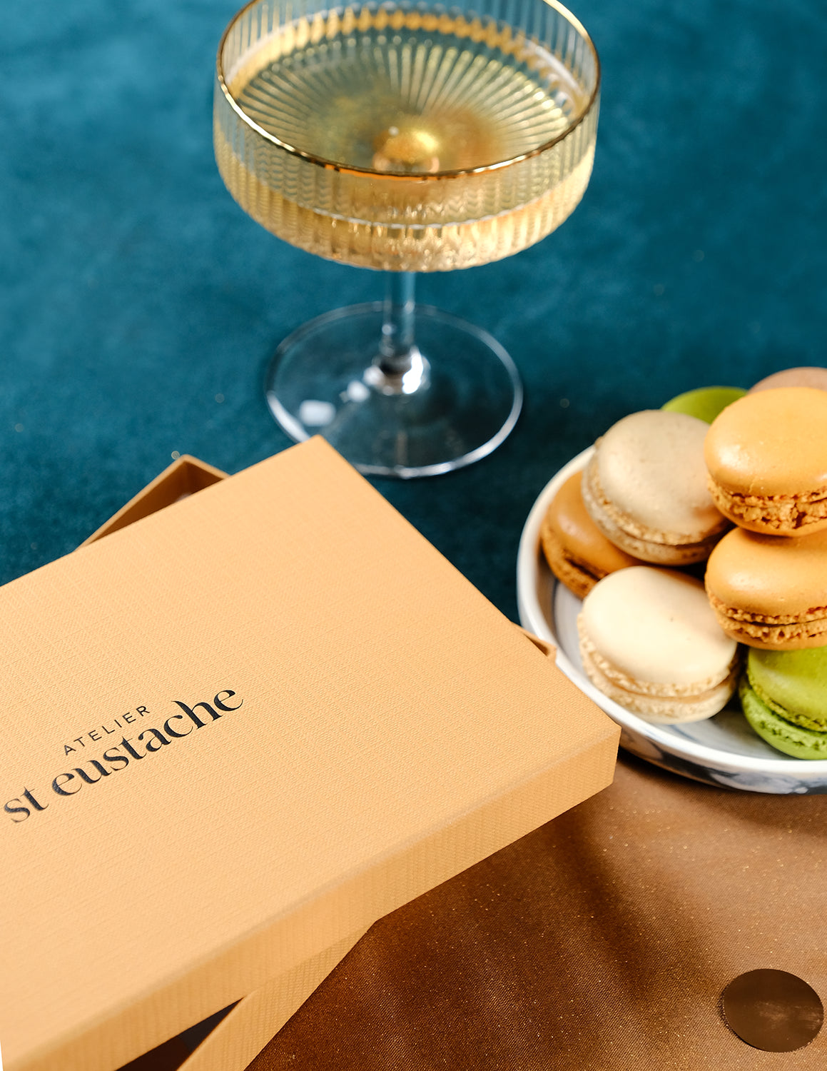 Coffret de Noël doré Atelier St Eustache à côté d&#39;une assiette de macarons et d&#39;une coupe de champagne, dans une ambiance festive et lumineuse pour Noël