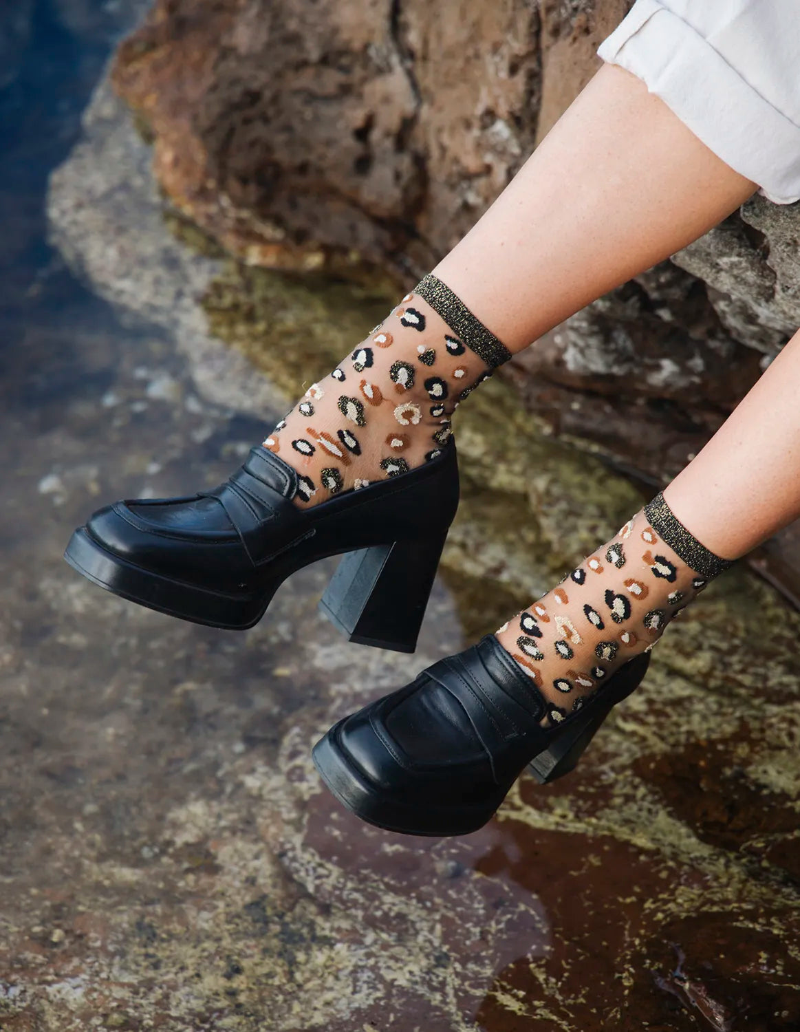 Zoom de la paire de chaussettes transparentes à motif léopard Horseshoe Bend Gold Atelier St Eustache portée par une mannequin assise au bord de l&#39;eau avec un des chaussures à talons noires