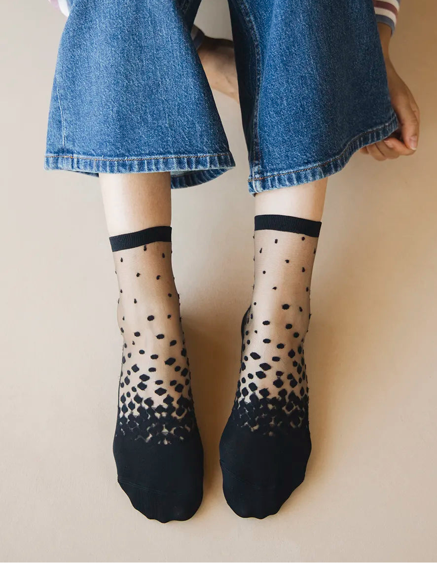 Chaussettes transparentes à pois noirs, portées par une femme assise par terre et portant un jean bleu