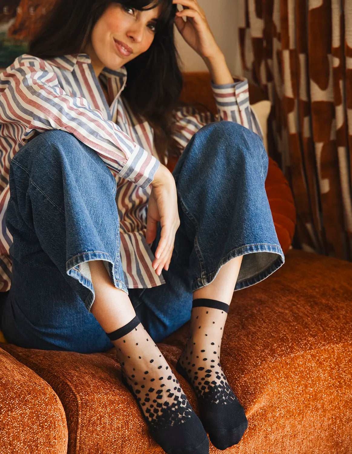 Chaussettes transparentes à pois noirs, portées par une femme assise sur un canapé portant un jean bleu et une chemise rayée