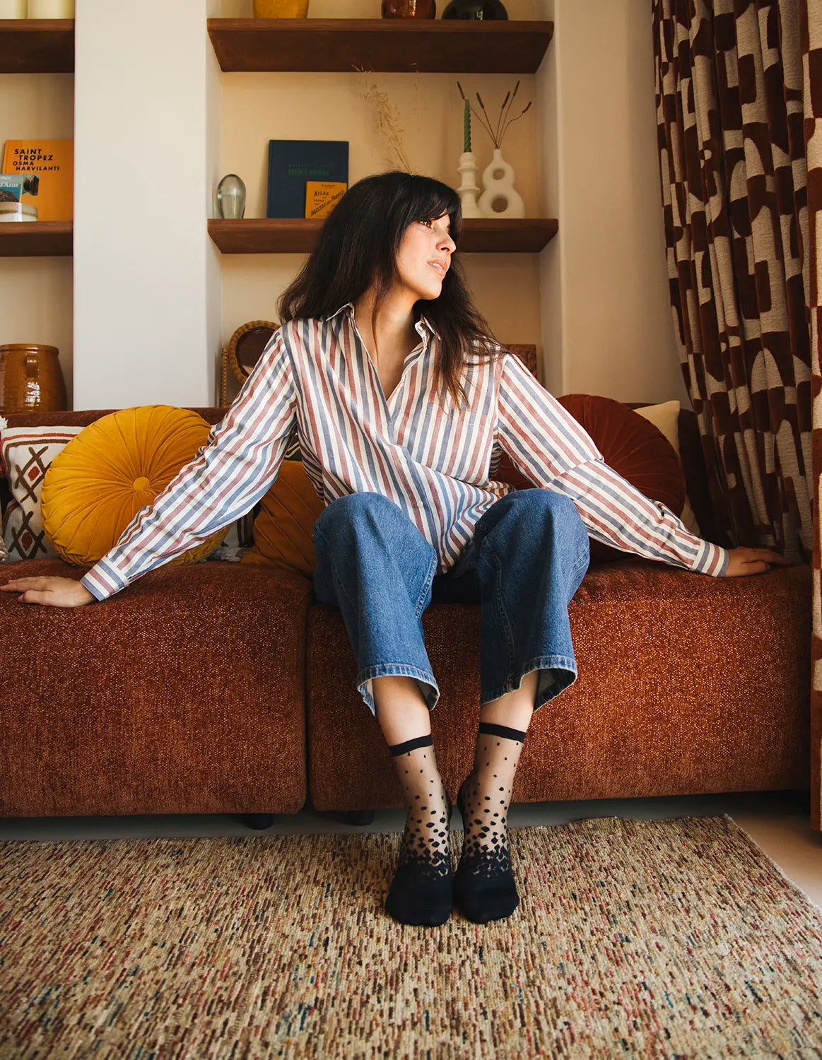 Chaussettes transparentes à pois noirs, portées par une femme assise sur un canapé portant un jean bleu et une chemise rayée dans une architecture moderne et 70&#39;s