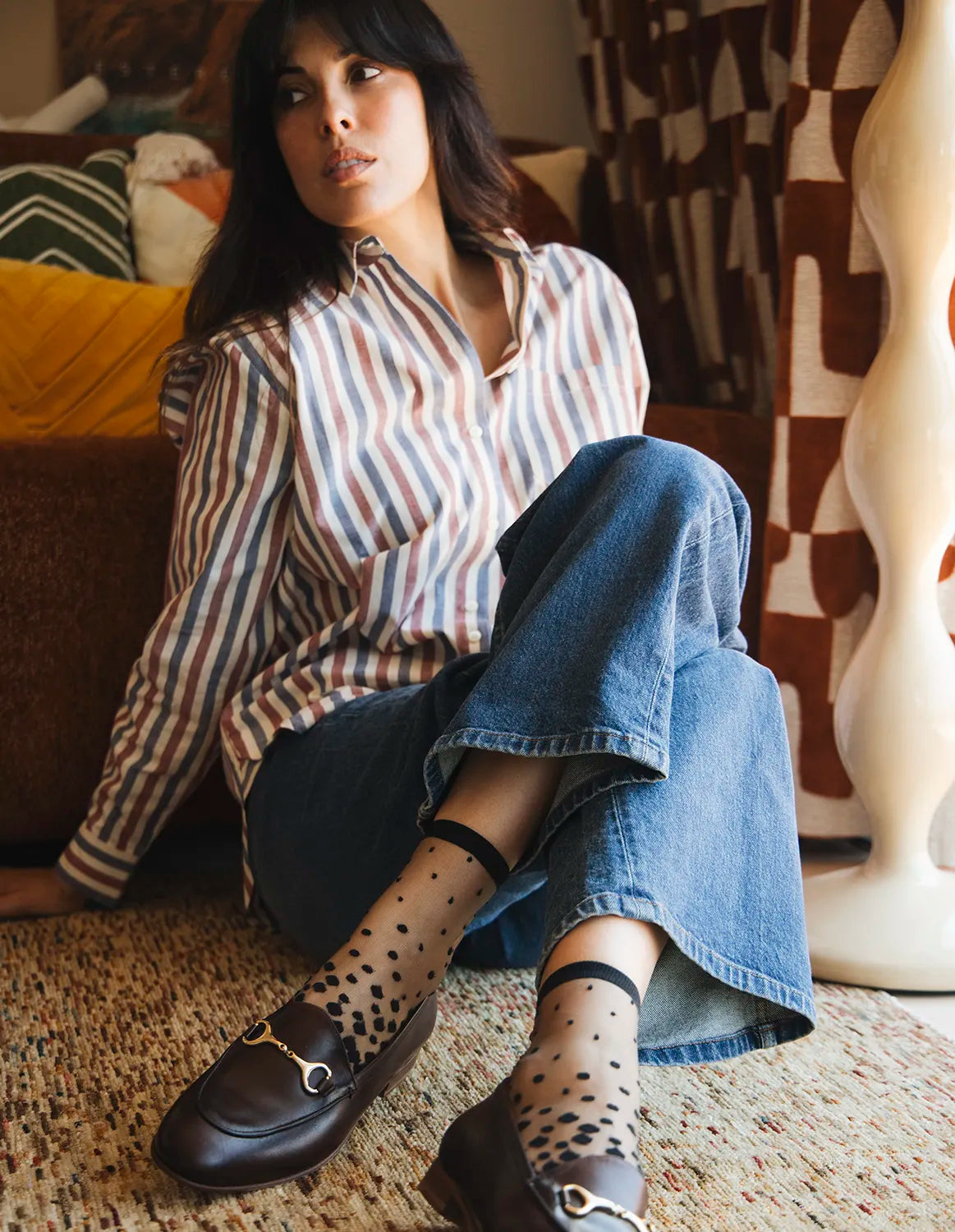 Chaussettes transparentes à pois noirs, portées par une femme assise par terre portant un jean bleu, une chemise rayée et des mocassins bordeaux
